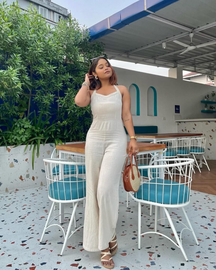 Woman in white outfit standing by outdoor cafe, holding a brown bag, with green plants and blue seating in the background.