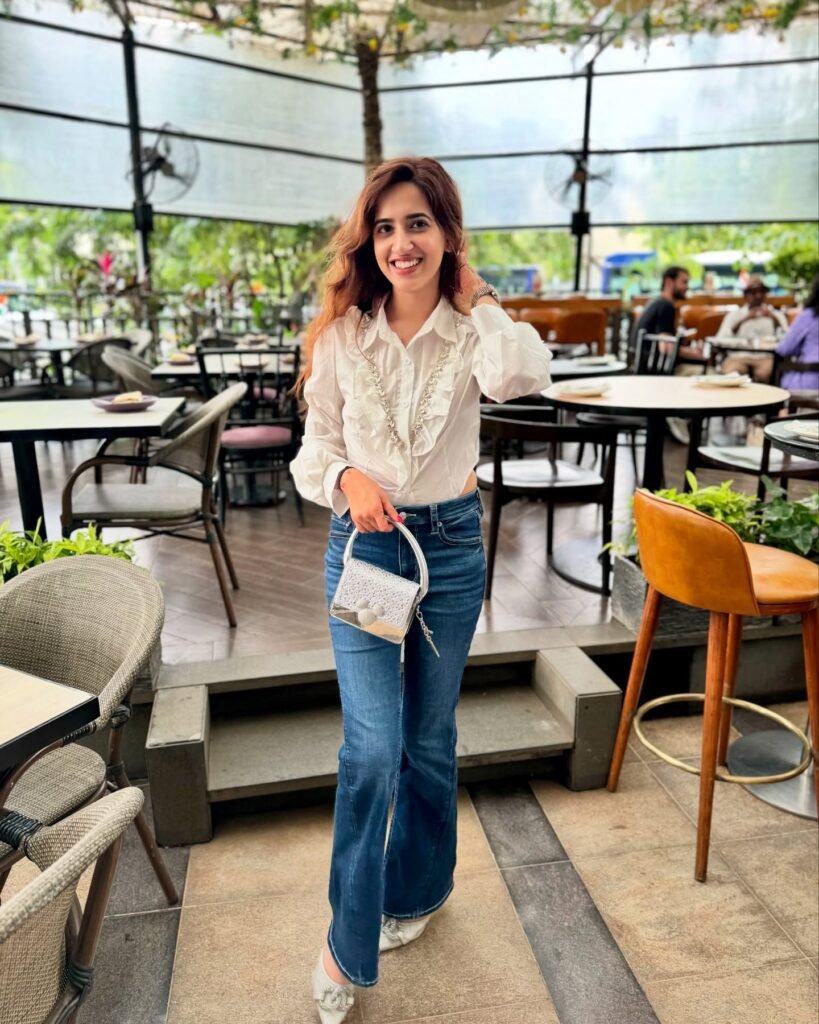Smiling woman in casual outfit with jeans and white blouse, holding a small bag in a stylish cafe setting.