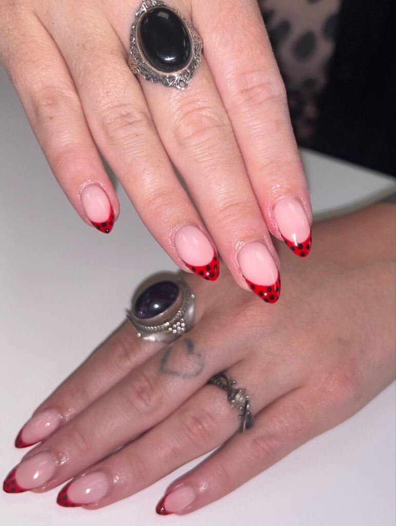 Close-up of elegant ladybug-inspired nail art with red tips and black dots, adorned with vintage rings.