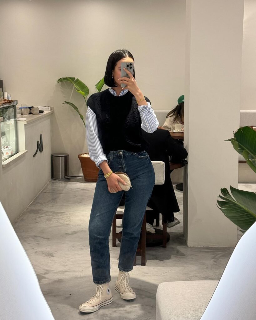 Casual cafe outfit with blue jeans, black sweater vest, striped shirt, and white sneakers, holding phone for selfie.