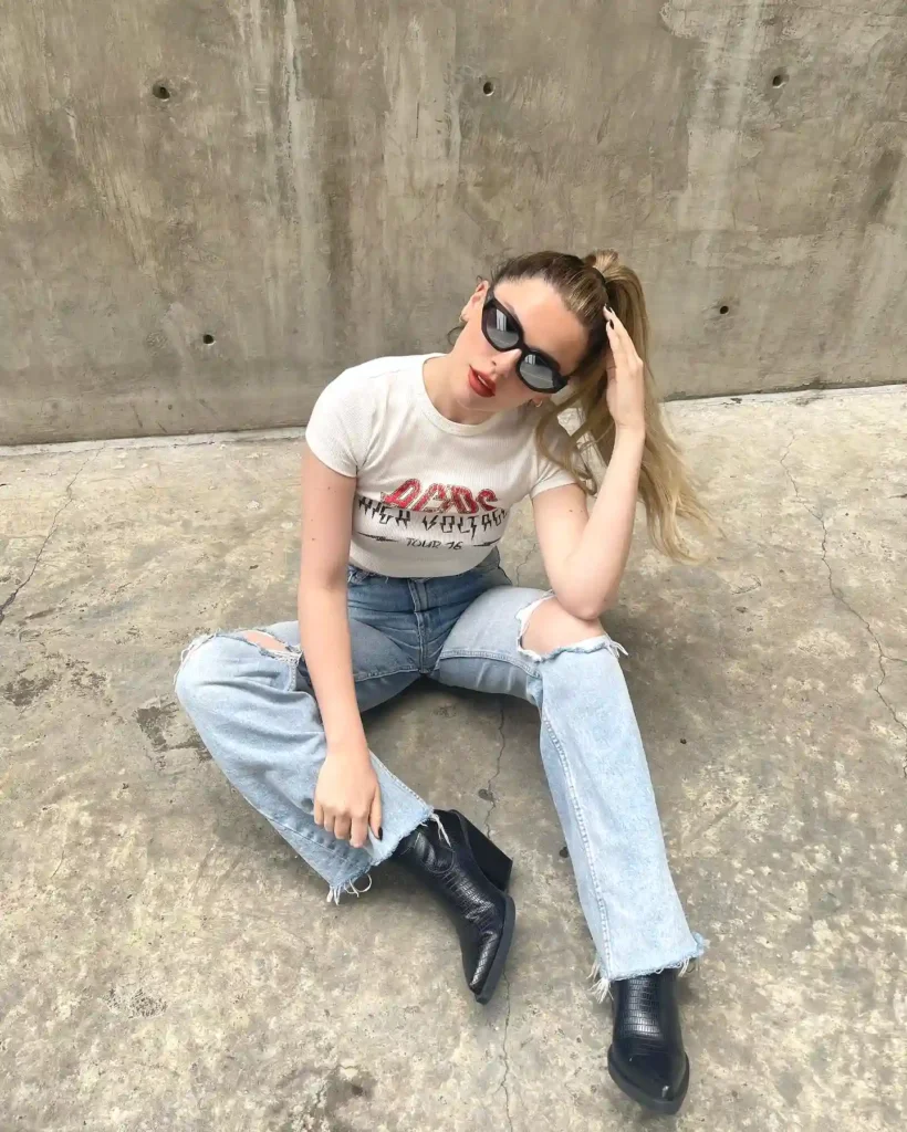 Young woman in sunglasses and casual outfit sitting on concrete floor with hand in hair.