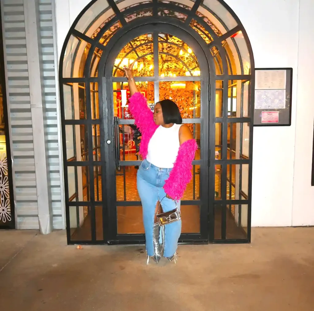Fashionable woman poses in pink jacket and jeans in front of an illuminated arched doorway.