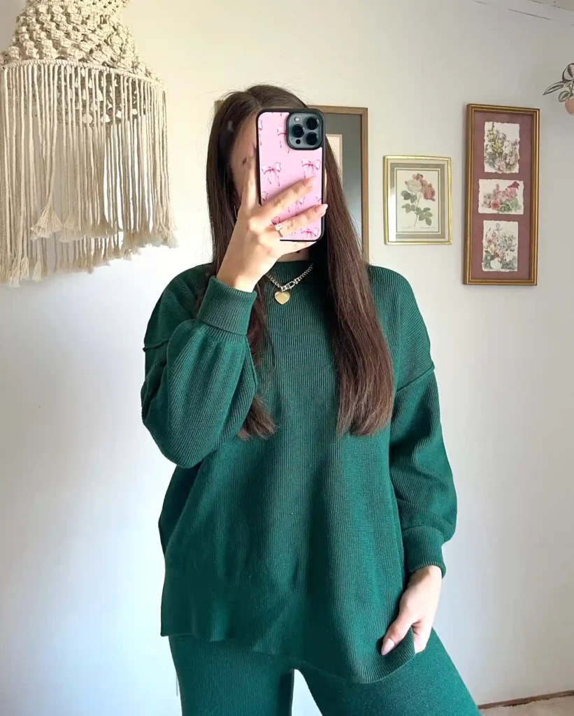 Person in green sweater taking a mirror selfie with a pink phone case, room decor in the background.