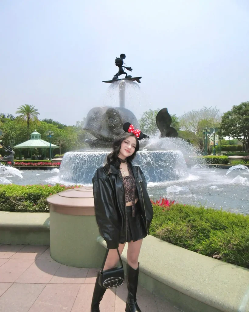 Young woman in stylish outfit poses by fountain with Mickey Mouse statue, sunny day at theme park.