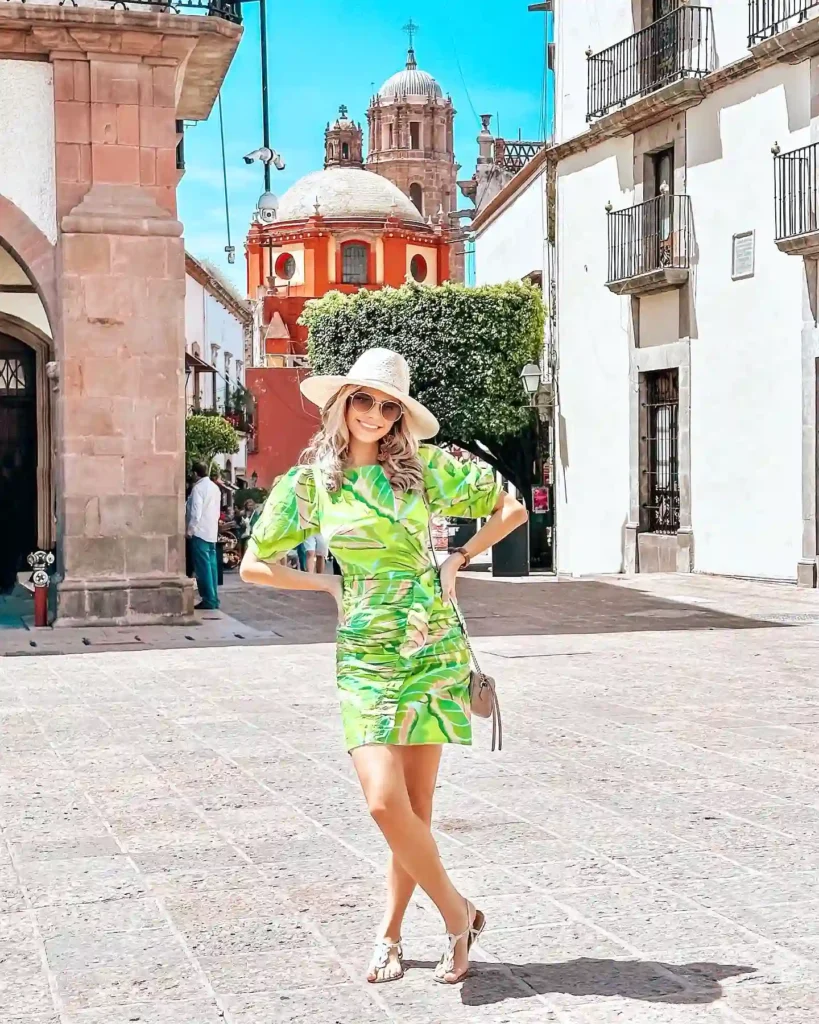 Woman in vibrant dress and hat poses in a sunny, historic town square with charming architecture.