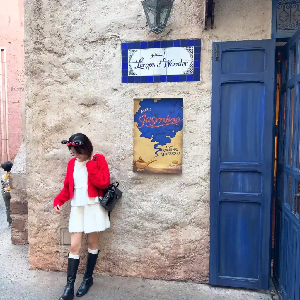 Woman in red jacket standing by Lamps of Wonder sign with Meet Jasmine poster in Moroccan-themed setting.