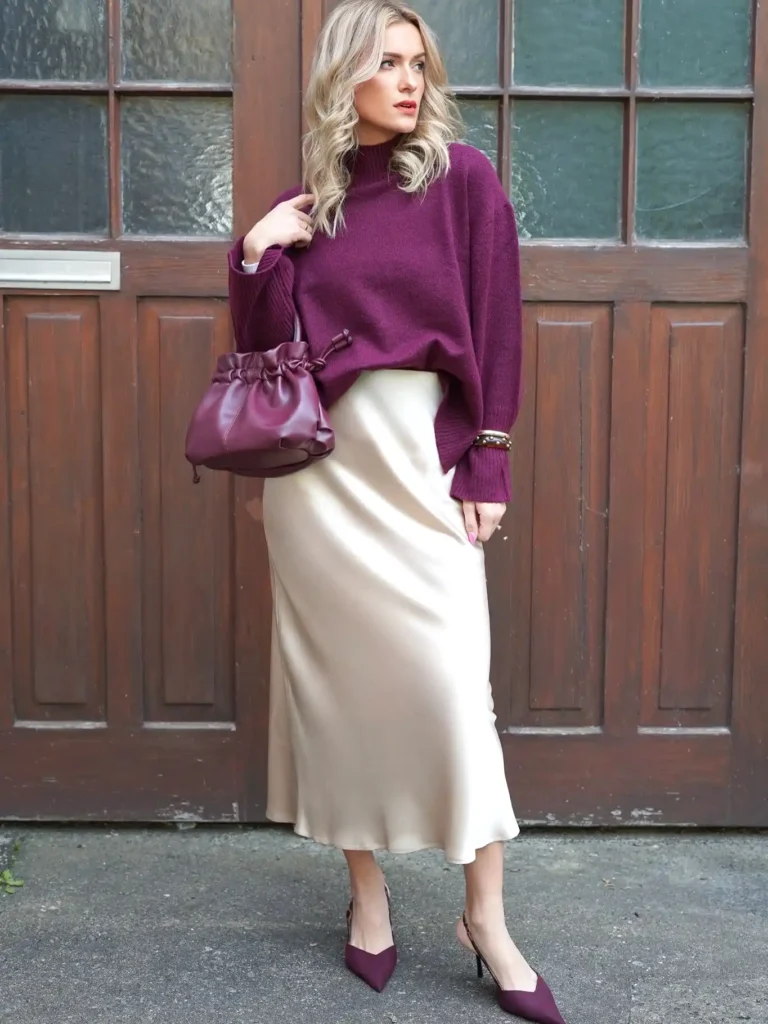 Woman in purple sweater and cream skirt holds matching bag in front of wooden door.