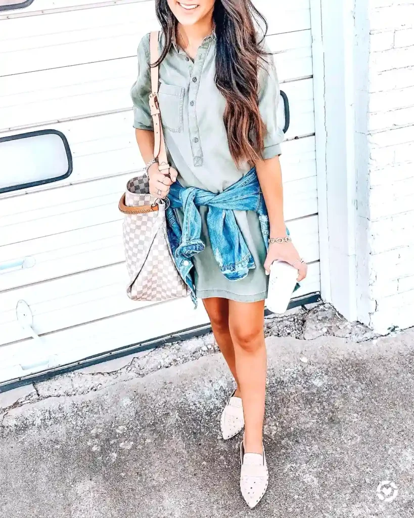 Woman in casual dress with denim jacket, designer bag, and a coffee cup standing outdoors against a white door.