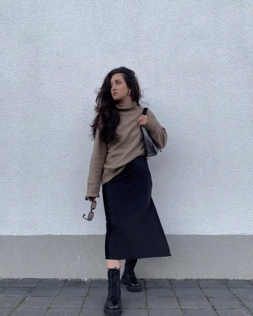 Woman in a stylish outfit with a sweater and skirt, holding sunglasses and a bag, standing against a white wall.