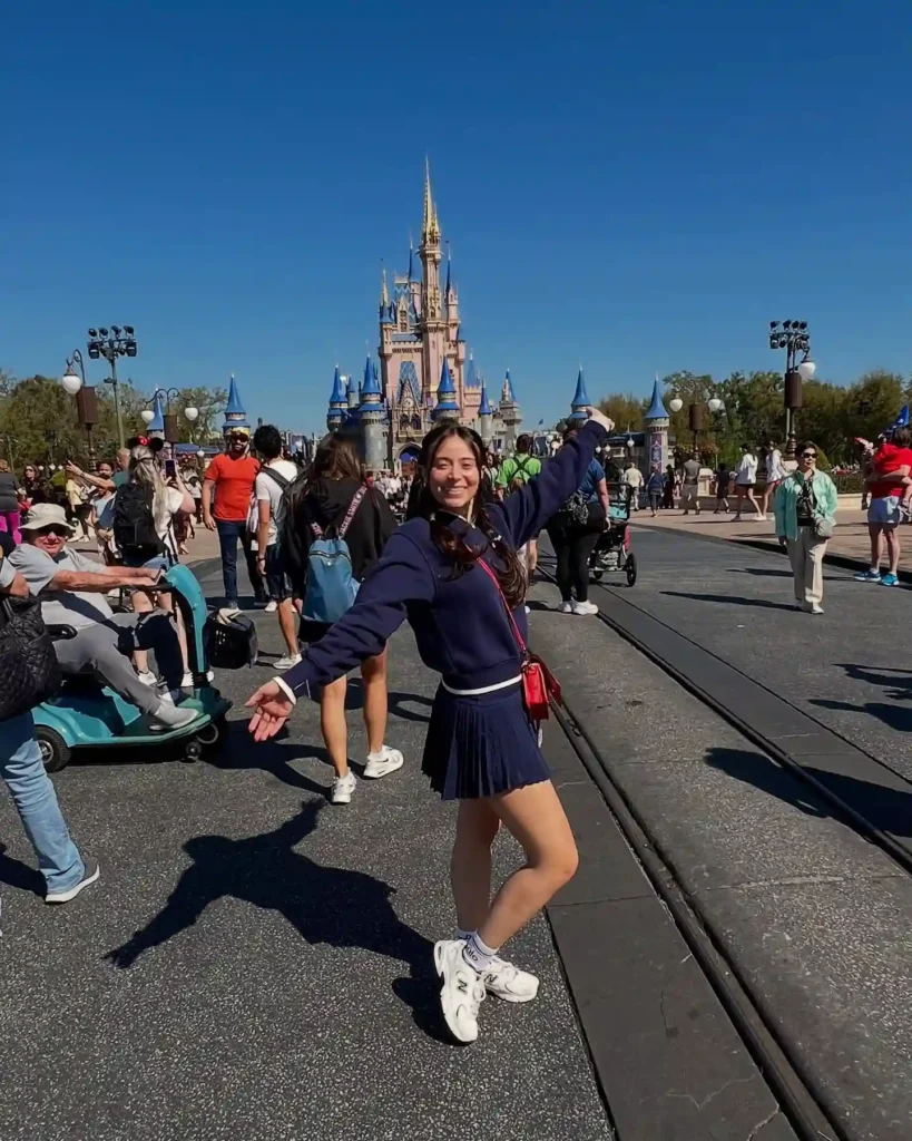 Woman joyfully posing in front of a castle at a theme park, surrounded by visitors under a clear blue sky.