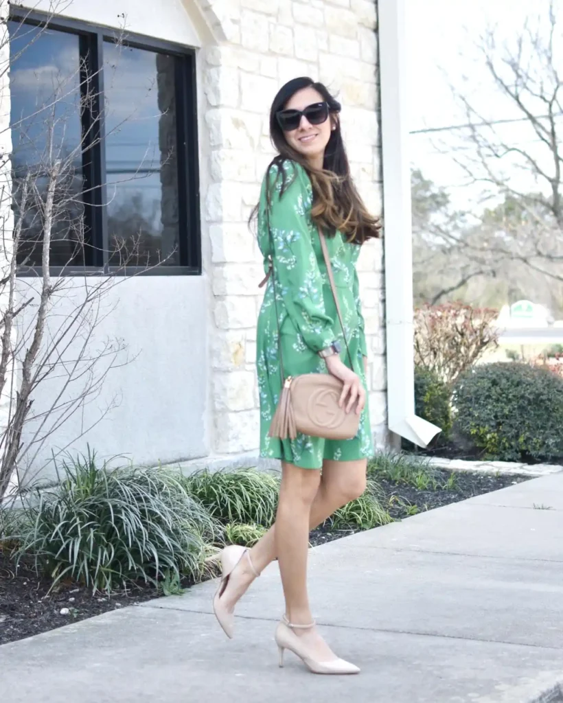 Woman in green floral dress with beige handbag and heels walking outside a stone building.