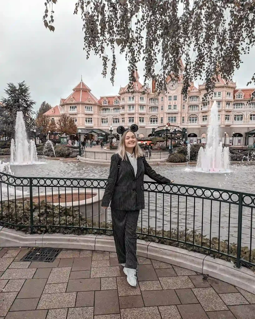 Person in a striped suit standing by a fountain, elegant pink building in the background, smiling under cloudy sky.