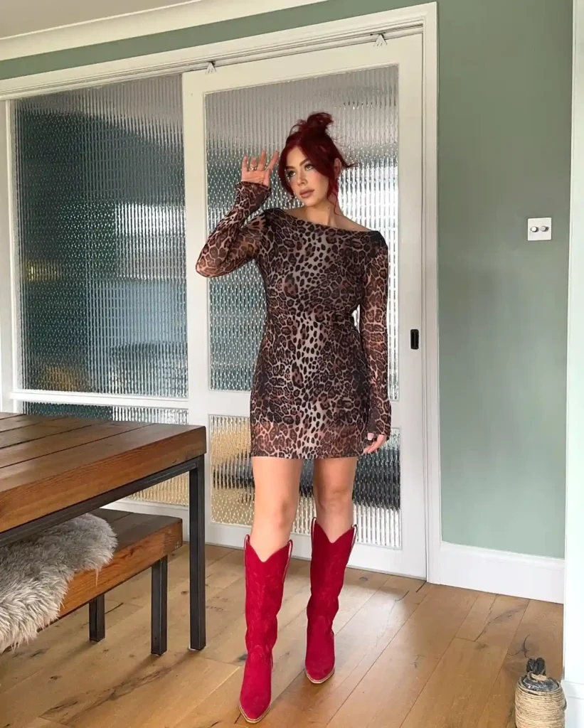 Red-haired woman in leopard print dress and red boots posing indoors next to a wooden table.