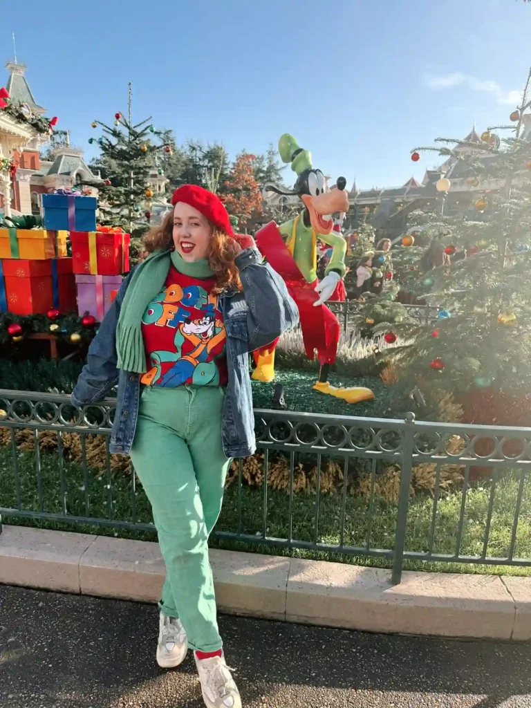 Smiling woman in festive outfit with holiday decorations and cartoon character at a theme park.