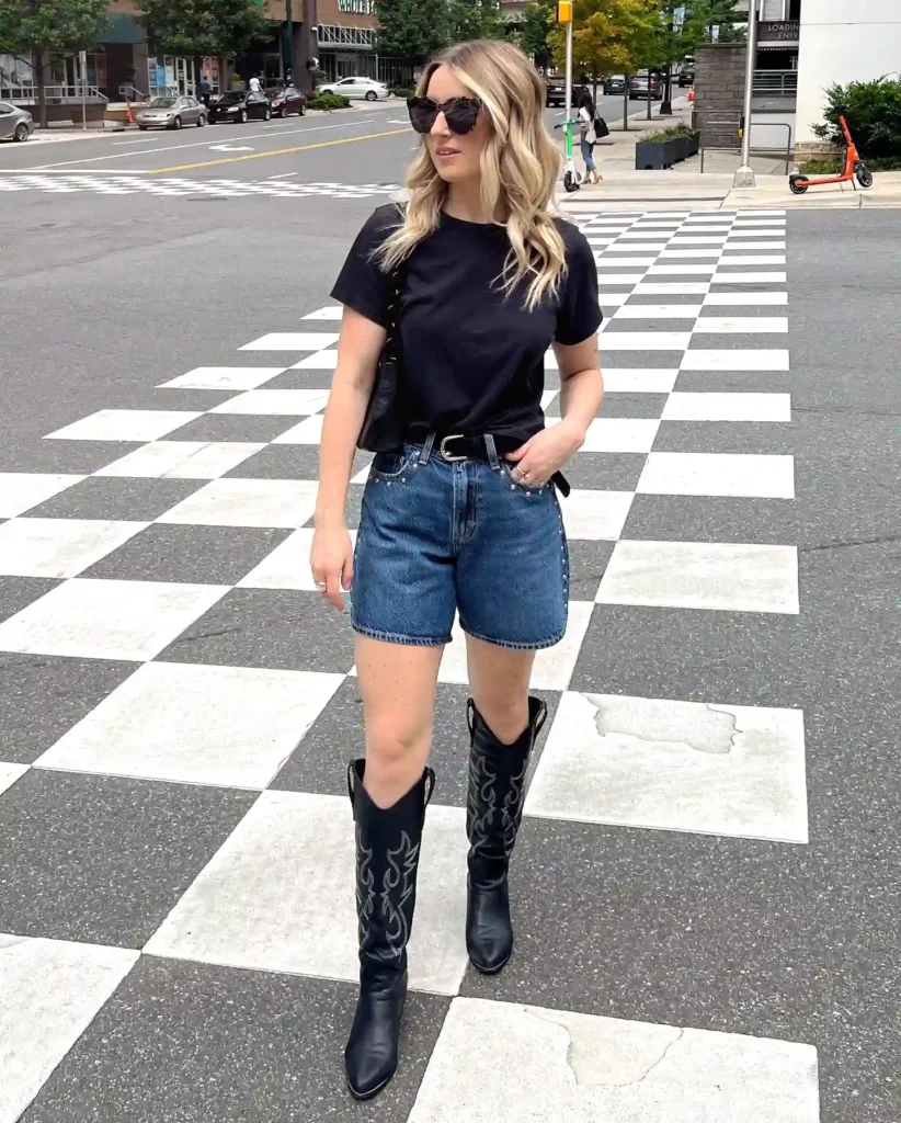 Woman in black shirt and denim shorts crossing a checkered street, wearing sunglasses and cowboy boots.