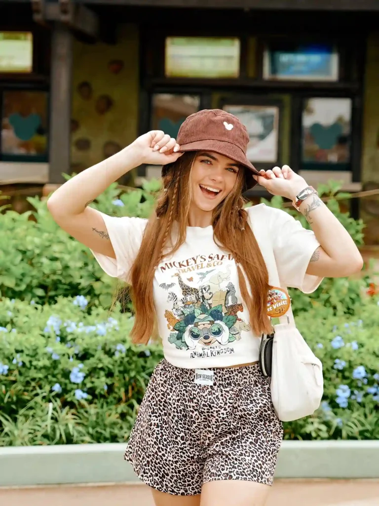 Smiling woman in a safari-themed shirt and bucket hat, standing outdoors with flowers in the background.