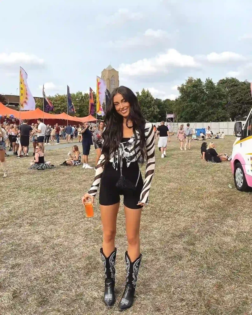 Festival-goer in stylish outfit and boots enjoys outdoor event with flags, holding a drink on a sunny day.