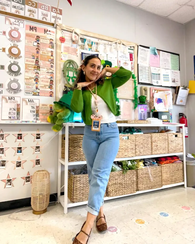 Smiling woman poses in a colorful classroom, wearing a green cardigan and jeans, surrounded by educational displays.
