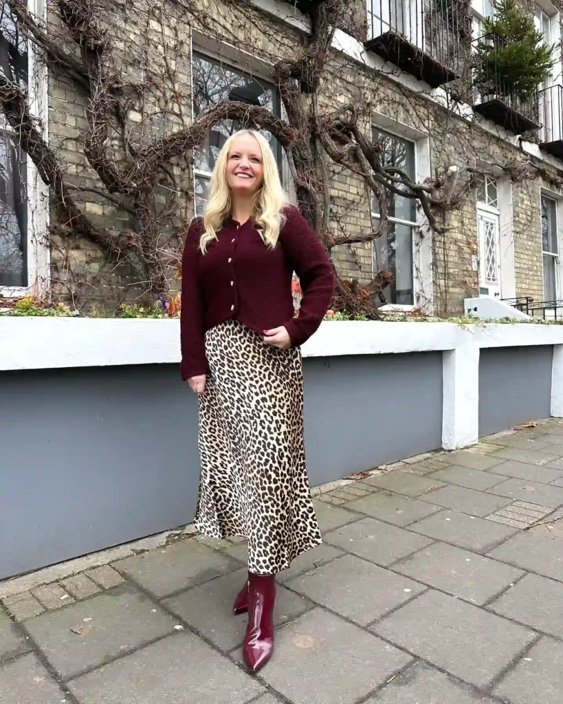 Woman in burgundy sweater and leopard print skirt smiling outside a vine-covered building.