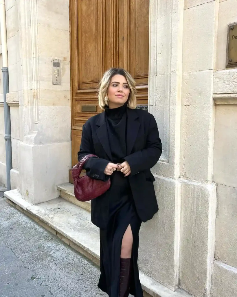 Woman in chic black outfit with burgundy bag stands by wooden door on city street.