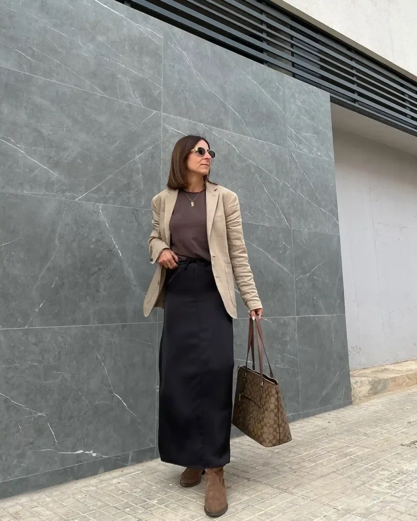 Woman in stylish beige blazer and black skirt, holding a tote bag, standing against a modern gray marble wall.