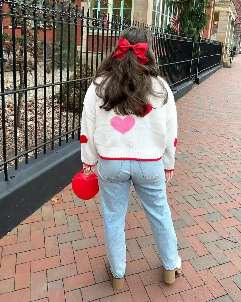 Woman with heart-patterned sweater and red bow walking on brick path, holding red heart-shaped purse.