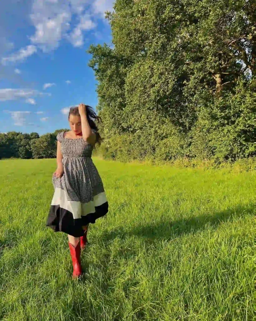 Woman in checked dress and red boots walking in sunny green field with blue sky and trees.