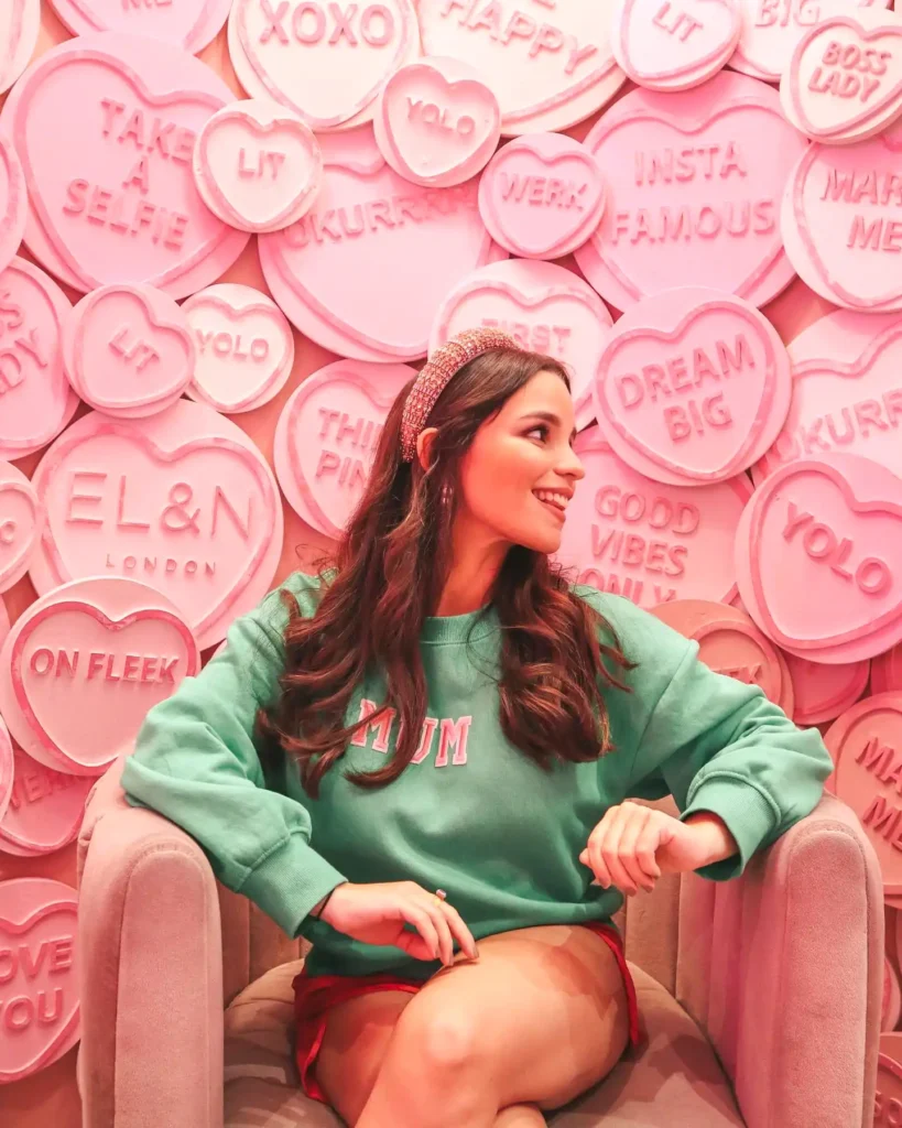 Woman smiling in a pink heart-themed cafe, wearing a green sweater with MUM text, surrounded by motivational phrases.