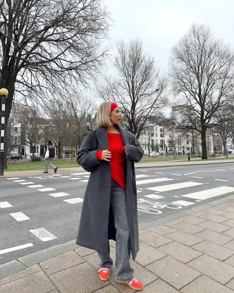 Woman in gray coat and red top crossing the street with bare trees and buildings in the background.