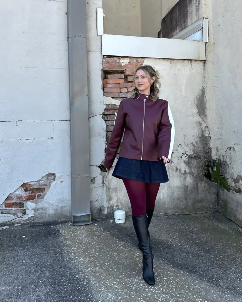 Fashionable woman in a burgundy jacket and boots walking by an old brick wall.