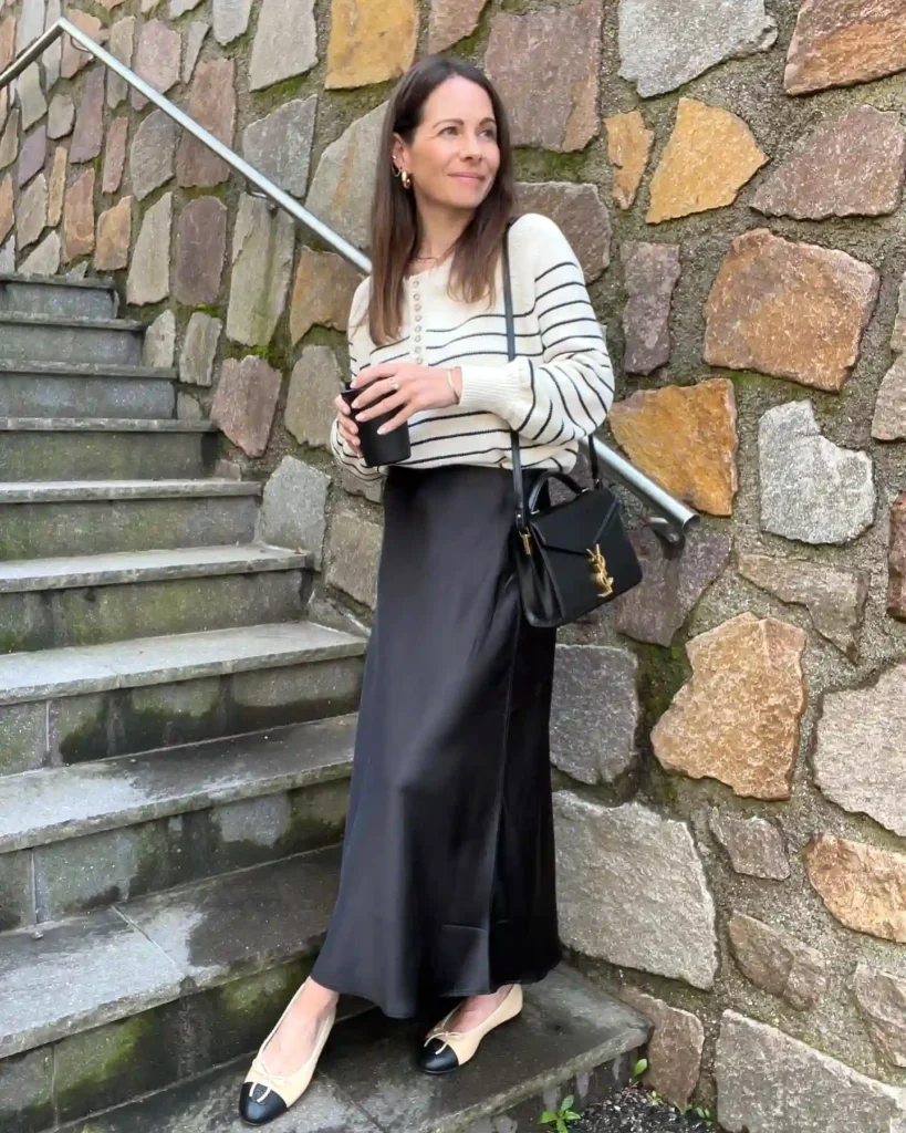 Woman in striped sweater and black skirt, holding coffee, stands on stone steps, smiling casually.