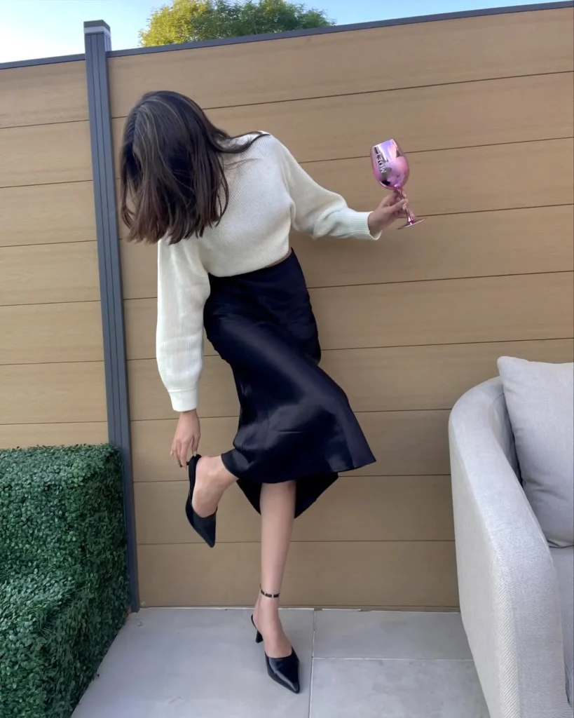 Woman adjusting high heel while holding a pink cup, wearing stylish outfit, standing outdoors by a wooden fence.