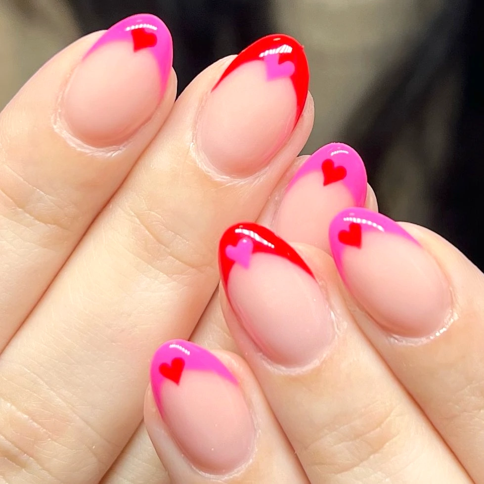 Almond-shaped nails with pink and red French tips and heart details, showcasing creative Valentine's nail art.