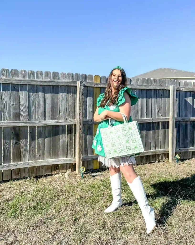 Woman in green dress and white boots smiles while holding a patterned tote bag in sunny backyard.