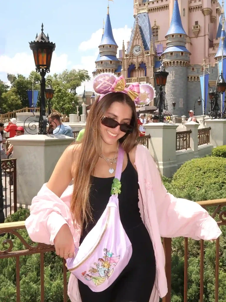 Smiling woman in front of a castle, wearing pink Minnie ears and a matching bag, at a theme park.