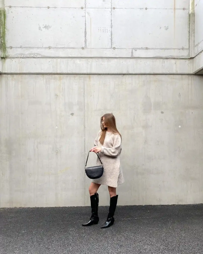 Woman in beige sweater dress and black boots against concrete wall, holding a black bag, outdoor fashion shot.