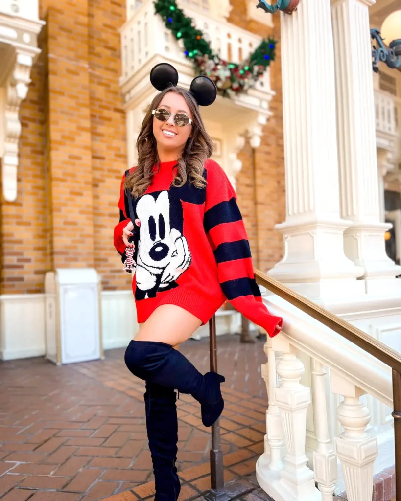 Woman in Mickey Mouse sweater and ears, smiling at theme park entrance with holiday decorations.
