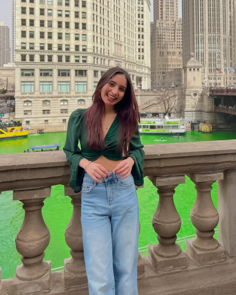 Young woman smiling by a river dyed green for St. Patrick's Day in a cityscape background.