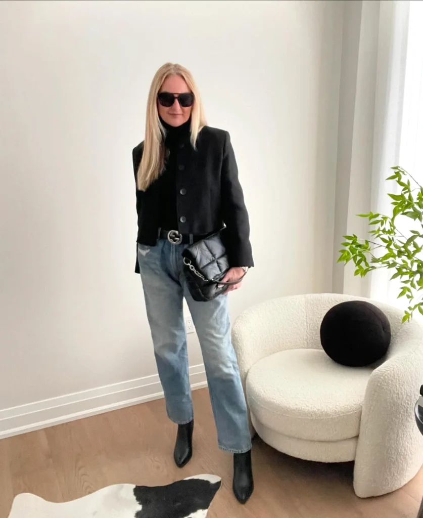 Stylish woman in sunglasses wearing black blazer and jeans, holding a quilted bag next to a modern chair and plant.