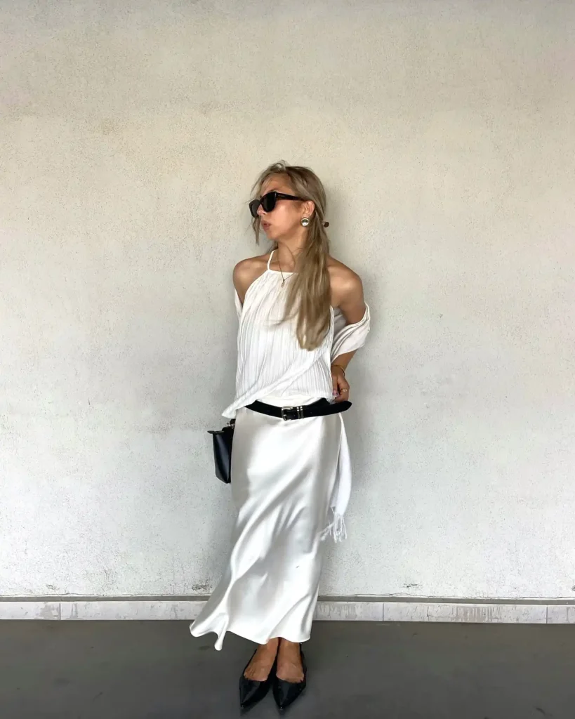 Stylish woman in white outfit poses with sunglasses and long skirt, against a neutral wall background. Fashionable and chic.