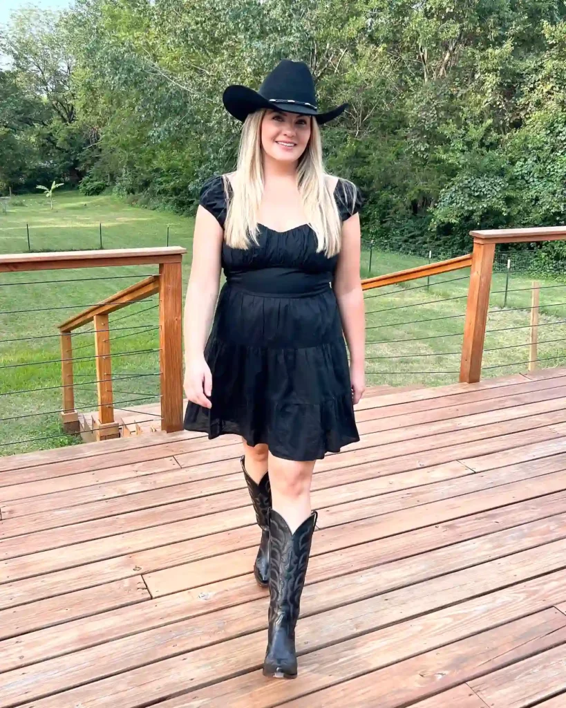 Woman in black dress and cowboy hat walking on a wooden deck with a scenic green backdrop.