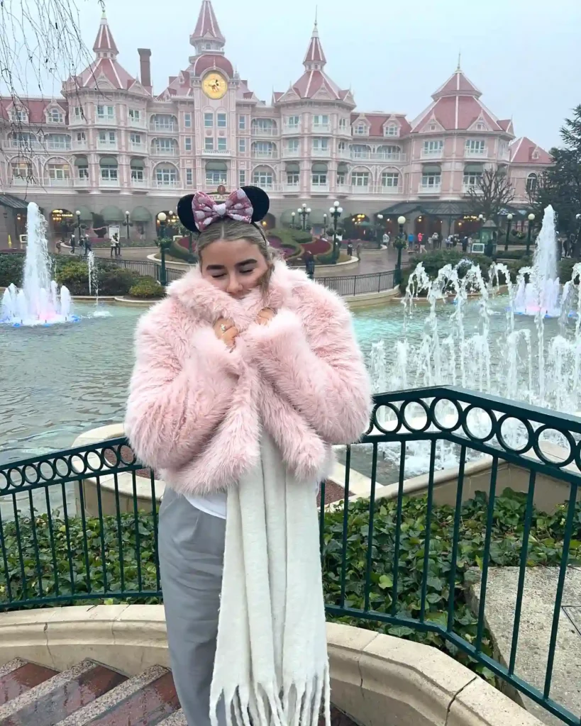 Woman in pink fur coat and Minnie ears at Disneyland entrance with fountains in the background.
