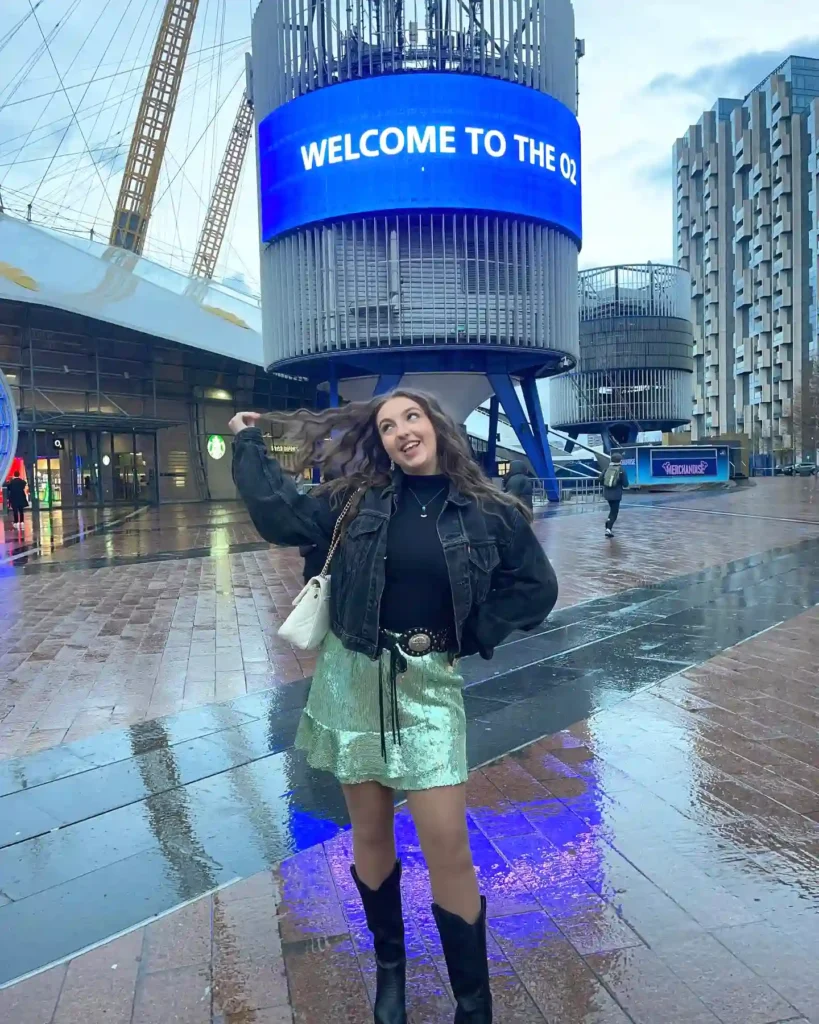 Happy woman posing outside The O2 arena in a stylish outfit, wet pavement reflecting blue digital signage.