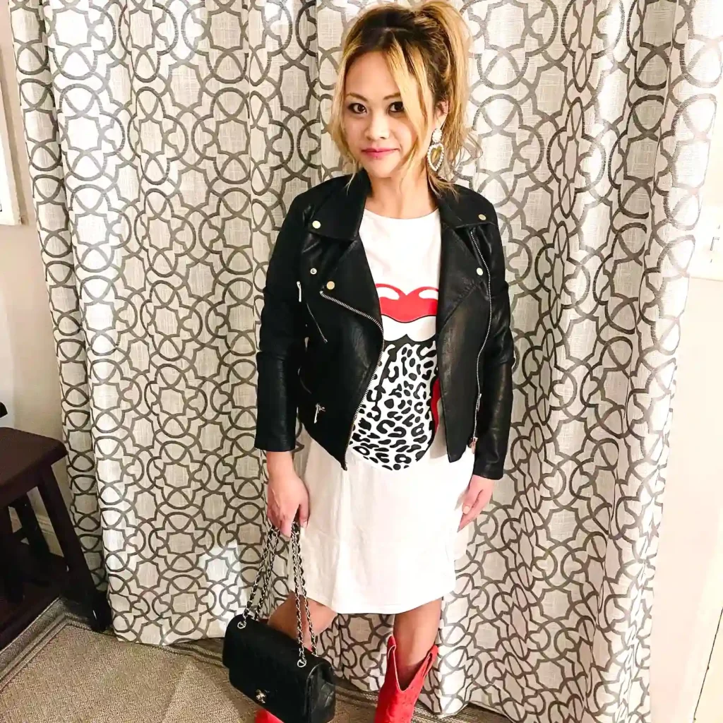 Woman in a leather jacket and red boots, holding a black purse, stands in front of patterned curtains.