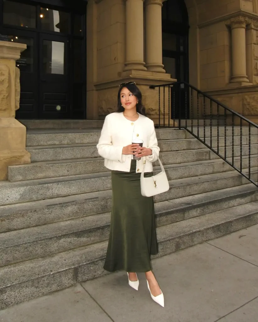 Woman in chic outfit with white jacket and green skirt holding a coffee on stone steps.
