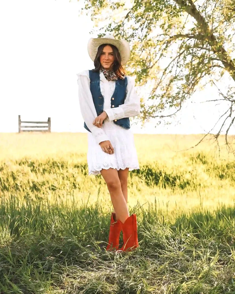 Cowgirl in red boots and white dress, denim vest, and hat stands in a sunny field by a tree.