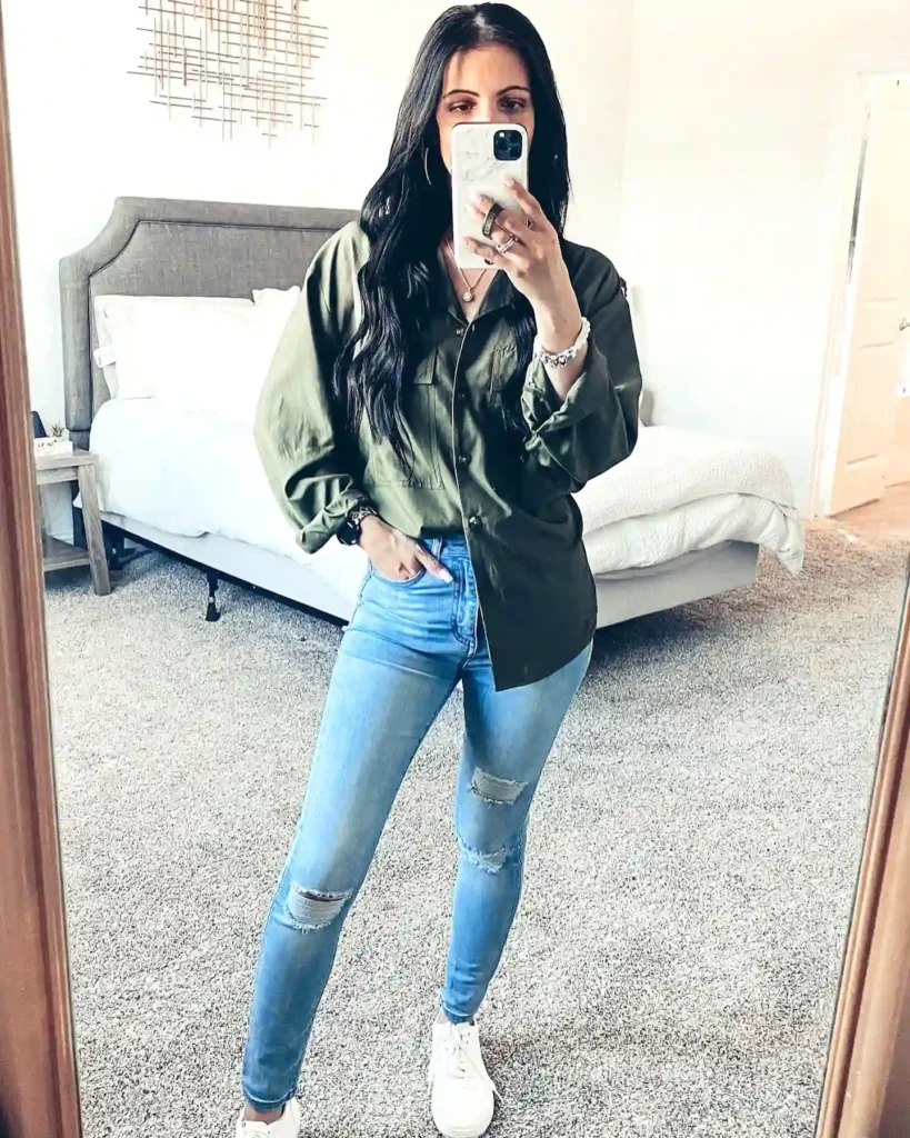 Person taking a mirror selfie wearing a green shirt and ripped jeans in a bedroom setting.