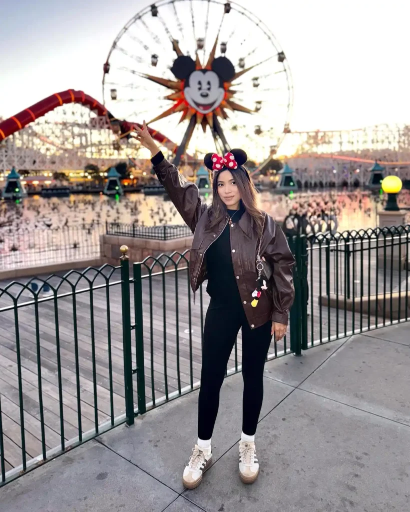Person posing in front of Mickey Mouse Ferris wheel at an amusement park, wearing Mickey ears and a brown jacket.