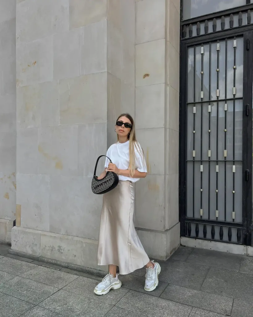 Stylish woman in white top and satin skirt, holding designer bag, wearing sneakers, stands by a stone building.