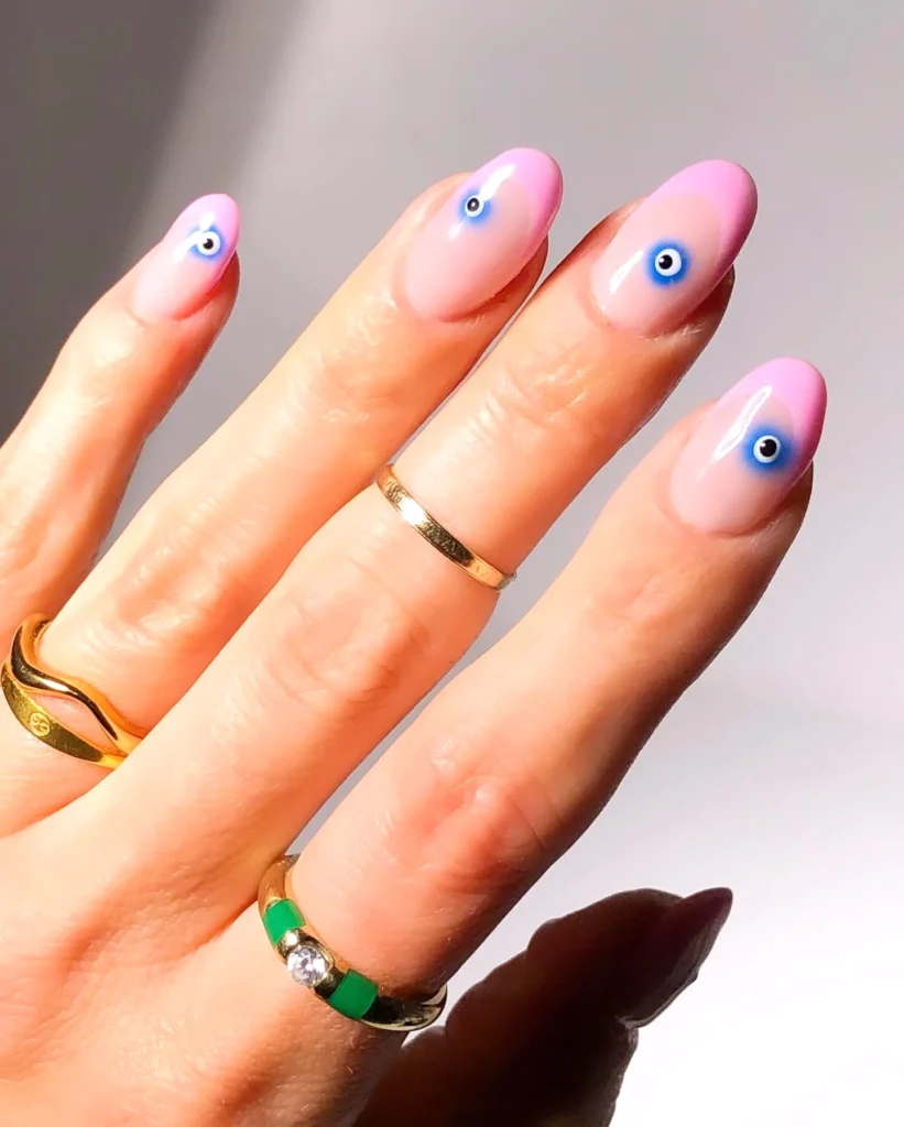 Hand with pink, evil eye nail art, wearing gold rings against a soft background. Trendy fashion nails.